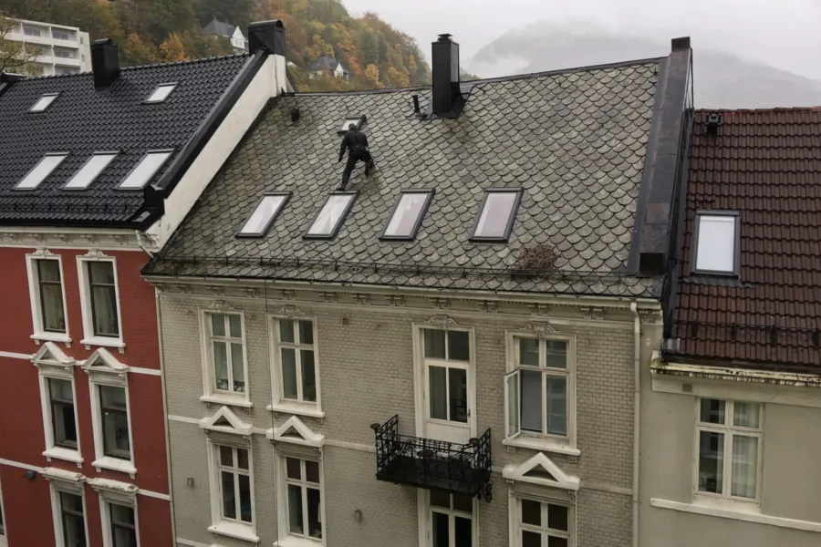 Dakdekker loopt over een grijs leien dak met dakramen om de schade te inspecteren, tussen hoge herenhuizen